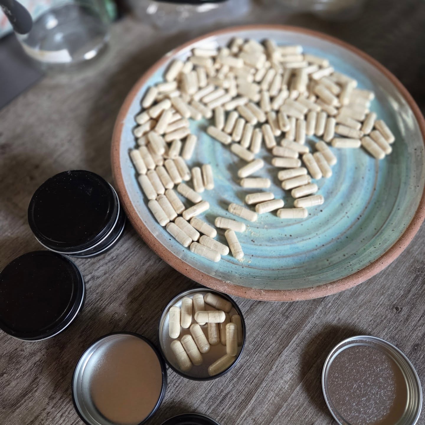 500770511_18507467290037498_4415060959033591650_n A close-up image of a handmade ceramic plate filled with beige capsules, surrounded by black containers, some of which are open revealing more capsules. The setting features a rustic wooden table, emphasizing the natural and organic aspect of the supplements.