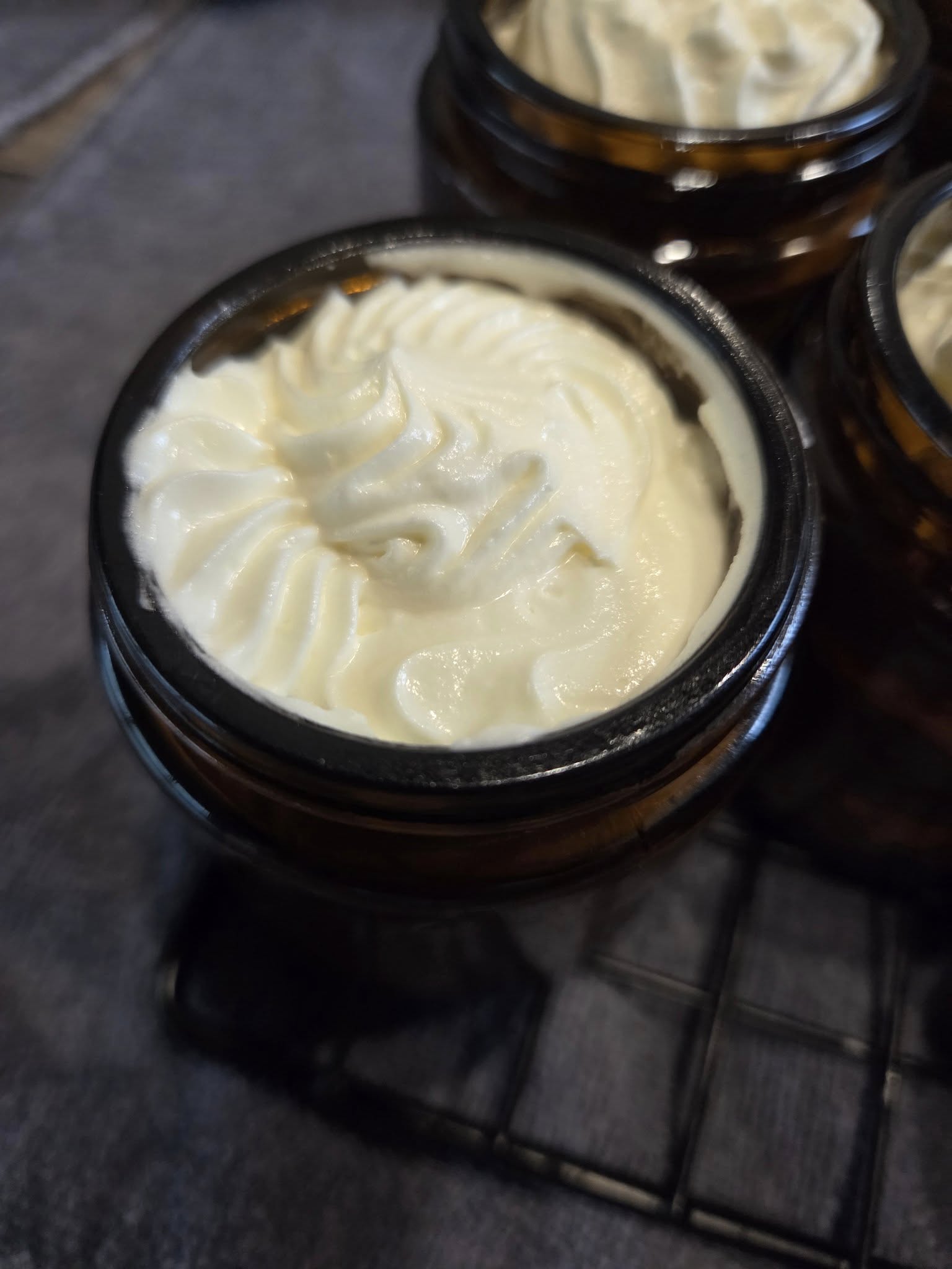 505890531_601474276312135_6899644611616977672_n Close-up of an amber glass jar filled with creamy, whipped body butter, showcasing a decorative swirl on top. The jar is placed on a black wire rack with a textured background, highlighting the product's luxurious appearance. Ideal for skincare enthusiasts looking for natural moisturizing solutions.