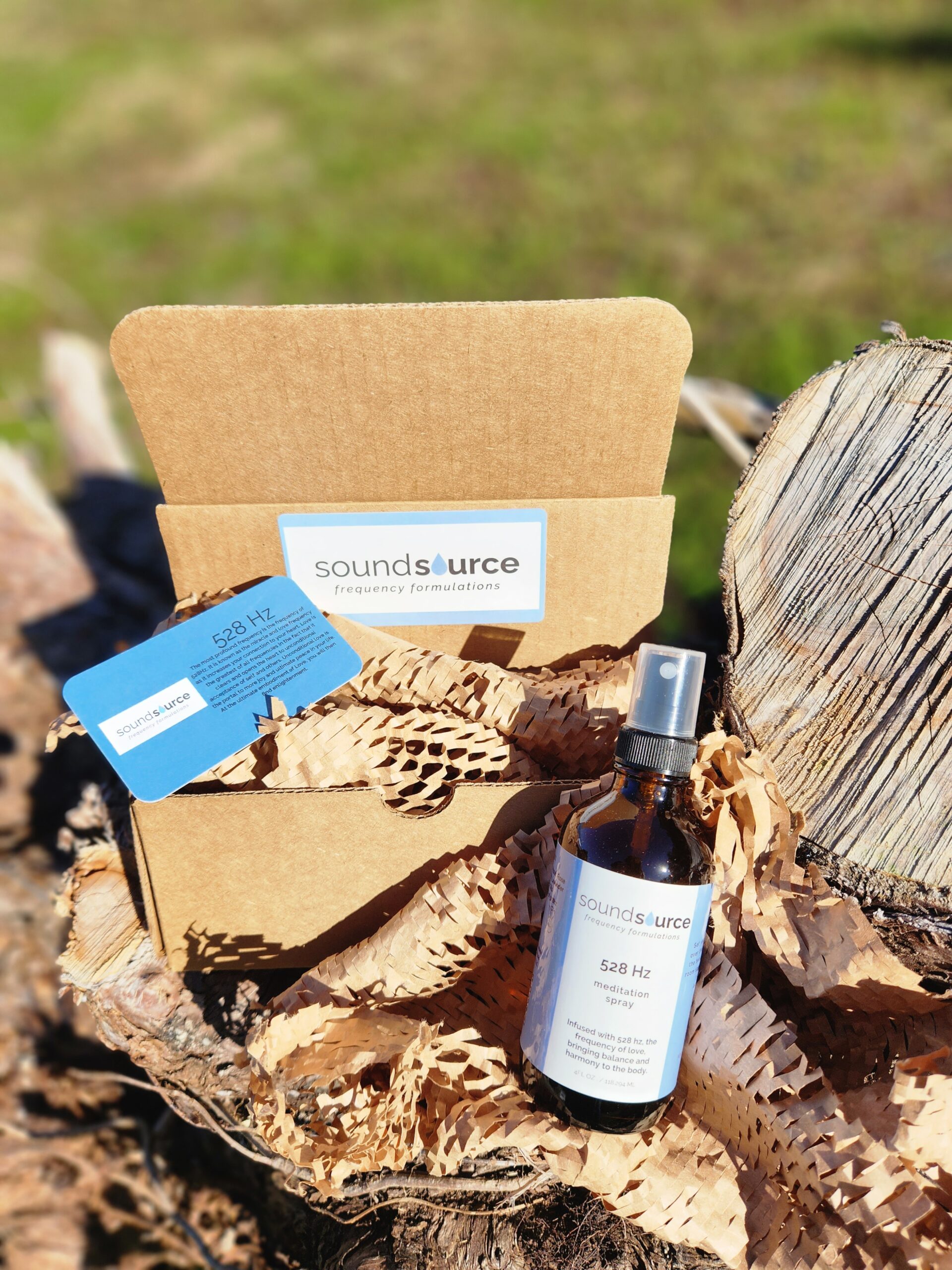 Caron Parker - 20230228_152019 Bottle of 528 Hz meditation spray from Soundsource, placed on a wooden surface with natural elements. The packaging includes a card and eco-friendly shredded paper, highlighting the product's focus on frequency formulations for meditation and wellness.